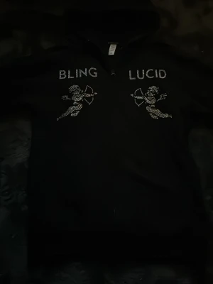 Svart hoodie från Bling Lucid - Säljer en svart hoodie från Bling Lucid med glittrande strasstenar som bildar texten 'BLING LUCID' och två änglar med pilbåge på bröstet. Hoodien har dragkedja och huva, tillverkad i mjuk bomullsblandning. Perfekt för dig som gillar streetwear med extra bling.