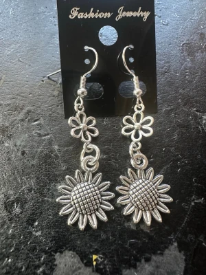 Silvriga solrosörhängen i metall - Unika örhängen i silverfärgad metall med solrosmotiv och små blommor ovanför. Hängande modell med krokfäste, perfekta för dig som gillar blommiga detaljer och vill sticka ut lite extra. Superfina att matcha med andra accessoarer i metall.