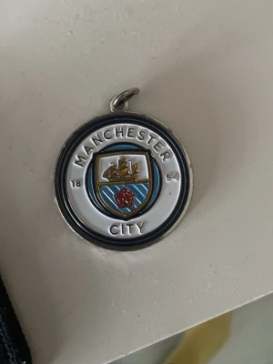 Manchester City halsbandshänge - Runt halsbandshänge i metall med Manchester Citys klubbmärke i blått, vitt, guld och rött emalj. Framsidan har klubbens logga och årtalet 1894, baksidan har en svart signatur. Perfekt för dig som älskar fotboll och vill visa ditt lag.