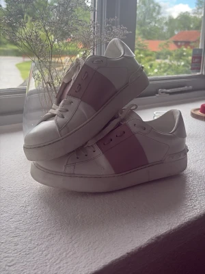 Valentino Garavani vita sneakers - Säljer ett par vita sneakers från Valentino Garavani med bred rosa läderdetalj över mitten. Använd inte alls många gånger. Klassisk låg modell med vita snören och diskret logga på plösen. Snygg och stilren design som passar till det mesta.