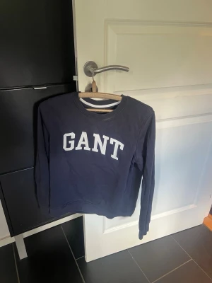 Mörkblå sweatshirt från GANT - Mörkblå sweatshirt från GANT med vit logotyp i college-stil över bröstet. Tröjan har rund halsringning och långa ärmar. Perfekt för en avslappnad och stilren look.