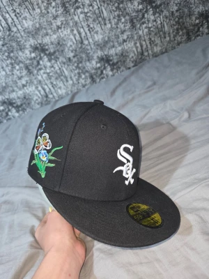 Svart New Era White Sox keps - Säljer en svart New Era 59Fifty keps med White Sox-logga i vitt framtill. Kepsen har broderade fjärilar och blommor i färgglada nyanser på sidorna, samt klassisk rak skärm. Perfekt för dig som vill sticka ut med snygga detaljer.