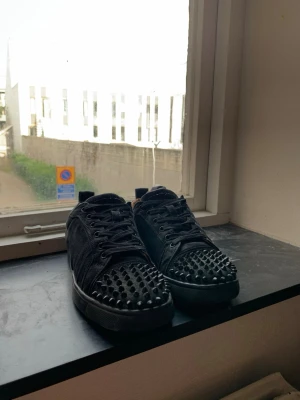 Svarta Christian Louboutin med nitar  - Svarta sneakers med coola nitar över tån och ovandelen. Skorna har snörning, platt sula och är tillverkade i mockaliknande material. Insidan är ljusbeige och de har en urban, edgy vibe som sticker ut i mängden.