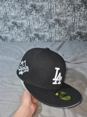 Svart LA Dodgers keps från New Era - Säljer en svart New Era 59FIFTY keps med LA Dodgers-logga i vitt framtill och World Series-märke på sidan. Kepsen har rak skärm och klassisk passform. Perfekt för dig som gillar streetwear och sportig stil.