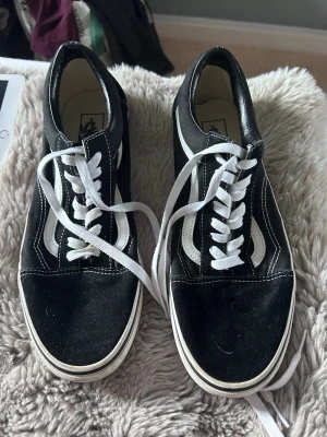Svarta Vans Old Skool sneakers - Klassiska Vans Old Skool sneakers i svart med vit sula och vita detaljer. Skorna har den ikoniska vita sidan, vita skosnören och kontrastsömmar. Ovandelen är i canvas och mocka, med lågt skaft för en avslappnad stil.