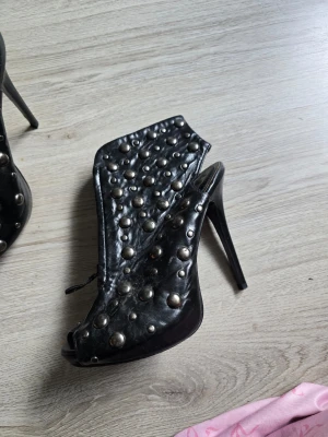 Svarta stövletter med nitar och öppen tå - Säljer ett par svarta stövletter med hög klack och öppen tå. Skorna är täckta med stora och små silverfärgade nitar för en edgy look. Dragkedja på insidan och platåsula framtill. Tillverkade i konstläder.