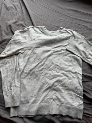Ljusgrå stickad tröja från H&M - En stilren ljusgrå stickad tröja från H&M med rund halsringning och ribbade muddar vid ärmslut och nederkant. Perfekt för lager-på-lager och enkel att matcha med olika outfits. Mjuk och tunn i materialet.