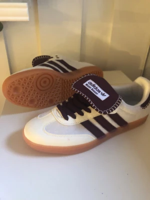 Adidas Samba Wales bonner OG vita och vinröda - Säljer ett par Adidas Samba OG sneakers i vitt och vinrött med klassiska tre ränder på sidan. Skorna har brun gummisula, vinröda skosnören och detaljer samt stor plös med Adidas-logga. Materialet är en mix av läder och mocka för en retrolook.