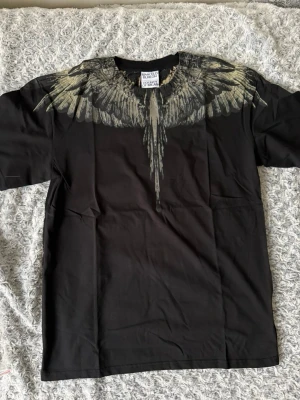 Svart t-shirt från Marcelo Burlon - Snygg svart t-shirt från Marcelo Burlon med ikoniskt vingtryck i grått och beige över axlar och bröst. T-shirten har rund hals och korta ärmar. Perfekt för dig som vill sticka ut med en unik design och streetwear-vibe.