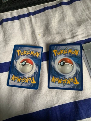 Två Pikachu Pokémonkort - Säljer två samlarkort med Pikachu från Pokémon. Ena kortet har en klassisk gul design med Pikachu i skogen och den andra har en modernare, färgstark illustration med Pikachu och en mindre Pikachu. Båda korten ligger i plastfickor.