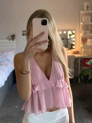 Ljusrosa volang topp med v-ringning - Söt ljusrosa topp med djup v-ringning och volangdetaljer. Toppen är ärmlös och har en luftig, croppad passform som ger en somrig känsla. Perfekt för dig som gillar en feminin och trendig stil.