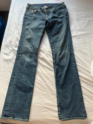 Blå bootcut jeans från True Religion - Säljer ett par blå bootcut jeans från True Religion med slitningar på låren och knäna. Klassiska fickor med vita sömmar och dekorativa knappar bak. Jeansen har låg midja och är gjorda i denim med en snygg tvätt.