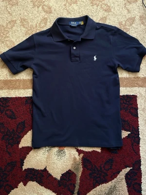 Mörkblå pikétröja från Ralph Lauren - Klassisk mörkblå pikétröja från Polo Ralph Lauren med vit broderad logga på bröstet. Tröjan har korta ärmar, krage och två knappar framtill. Tillverkad i bomull för en bekväm och stilren look.