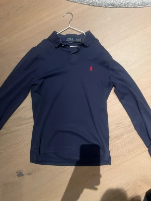 Mörkblå långärmad piké från Polo Ralph Lauren - Klassisk mörkblå långärmad pikétröja från Polo Ralph Lauren med röd broderad logga på bröstet. Skjortan har krage, knappar vid halsen och är tillverkad i mjuk bomull. Perfekt för en stilren och avslappnad look.
