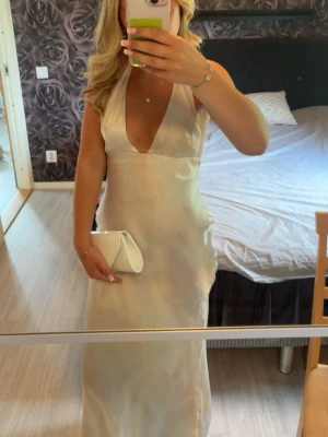 Champagnefärgad långklänning med djup v-ringning - Elegant långklänning i champagnefärgad satin med djup v-ringning och breda axelband. Klänningen har en figurnära passform och släpper mjukt längs kroppen. Perfekt för student eller bal. Matchas snyggt med en vit clutch.