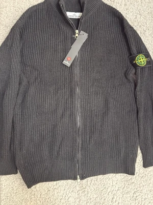 Svart stickad ziptröja Stone Island - Snygg svart stickad ziptröja från Stone Island med ribbad struktur och hög krage. Tröjan har dragkedja framtill och den klassiska Stone Island-loggan på vänster ärm. Perfekt för lager på lager och streetwear-stil.