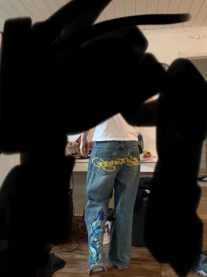 Ed Hardy jeans med drakbroderi - Säljer ett par blå jeans från Ed Hardy med fett broderad drake i blått, gult och svart längs ena benet. Baktill finns en stor gul Ed Hardy-logga. Modellen är loose fit med raka ben och klassisk femficksdesign. Perfekt för dig som vill sticka ut med coola detaljer. Notera ett hål vid backfickan se sista bilden, de märks knappt av vid andvändning.