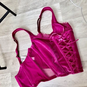 Rosa satin korsett från Victoria Secret  - Snygg rosa korsett i glansigt satinmaterial med spetskant upptill och dekorativa snörningar framtill. Justerbara axelband och hakknäppning bak. Perfekt för dig som vill ha en feminin och trendig look.