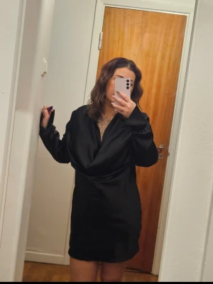 Svart Satinklänning  - Snygg svart kortklänning i satin med djup v-ringning. Klänningen har långa ärmar och en elegant, glansig finish som ger en lyxig känsla. Perfekt för dig som vill sticka ut med en stilren look.