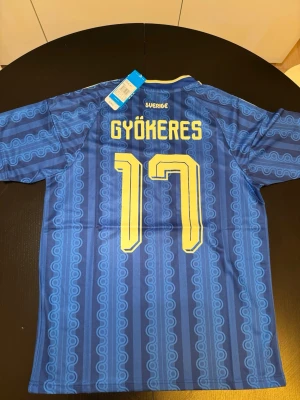 Blå Sverige fotbollströja Adidas - Snygg blå fotbollströja från Adidas med gula detaljer och trycket 'GYÖKERES 17' på ryggen. Tröjan har Sveriges landslagsemblem på bröstet och klassiska Adidas-ränder på axlarna. Tillverkad i lätt och ventilerande material, perfekt för match eller träning.
