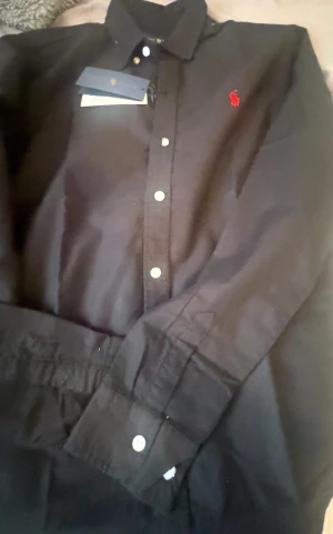 Svart skjorta från Polo Ralph Lauren - Svart skjorta från Polo Ralph Lauren med klassisk krage och knappar framtill. Skjortan har lång ärm och ett rött broderat logomärke på bröstet. 10/10 nytt skick 
