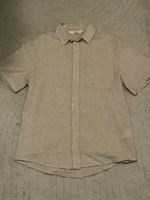 Beige linneskjorta från H&M - Säljer en stilren beige skjorta från H&M i linne. Skjortan har korta ärmar, klassisk krage, bröstficka och knappar framtill. Perfekt för varma dagar och enkel att matcha med olika outfits.