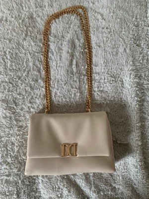 Beige handväska med guldkedja - Stilren beige handväska från Glitter med guldfärgad kedjerem och elegant D&D-logga framtill. Väskan har en mjuk, rektangulär form och är tillverkad i slätt konstläder. Perfekt accessoar för att lyfta din outfit. Har använt 2 gånger!