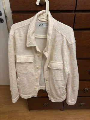 Krämvit overshirt från Zara - Stilren krämvit overshirt från Zara med två stora bröstfickor och knappar framtill. Overshirten har en avslappnad passform, klassisk krage och är tillverkad i ett mjukt, strukturerat tyg som ger en modern look.