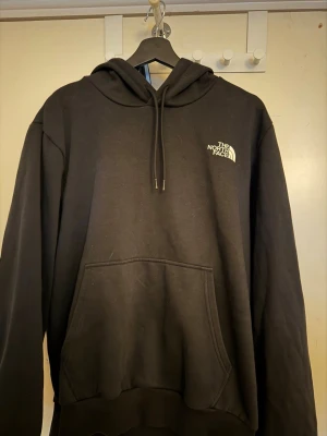 Svart hoodie från The North Face - Säljer en svart hoodie från The North Face med klassisk logga på bröstet och ryggen. Hoodien har huva med dragsko och en stor magficka framtill. Tillverkad i mjukt material som känns skönt mot huden. Perfekt för en avslappnad och stilren look.