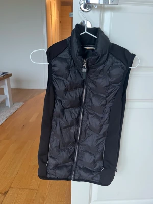 Svart dunväst med dragkedja - Snygg svart dunväst med hög krage och dragkedja framtill. Västen har quiltade partier på fram- och baksidan samt släta sidopaneler för en sportig look. Två fickor med dragkedja framtill. Perfekt för lager-på-lager och streetwear.