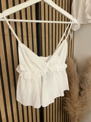 Vit linnetopp med volangdetalj - Söt vit linnetopp från Stradivarius med tunna axelband och volangdetalj under bysten. Toppen har en luftig och lätt känsla, perfekt för varma dagar. Materialet är mjukt och toppen har en v-ringad skärning framtill.