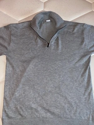 Grå stickad halvzip tröja - Snygg grå stickad tröja med halvzip och hög krage. Tröjan har ribbade muddar vid ärmslut och nederkant, perfekt för lager på lager. Enkel och stilren design utan synligt märke. Passar till många olika outfits. Använd en gång för skolfoto sen dess har jag aldrig använt den.