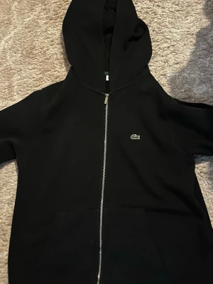 Svart zip hoodie från Lacoste - Säljer en svart hoodie från Lacoste med dragkedja framtill och klassisk krokodillogga på bröstet. Hoodien har huva och stora fickor framtill. Perfekt för en chill och stilren look.