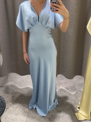 Ljusblå Balklänning/Satin klänning - Elegant ljusblå långklänning med v-ringad hals och breda, korta ärmar. Klänningen har markerad midja och är tillverkad i ett glansigt satinliknande material som ger en lyxig känsla. Perfekt för dig som vill sticka ut med en stilren och modern look. Från Gina Tricot och helt oanvänd. Storlek 36, är 160 som referens och den är lite lång som syns på andra bilden. Helt slutsåld på hemsidan