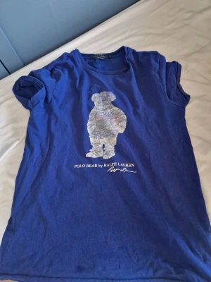 Blå Polo Bear t-shirt från Ralph Lauren - Blå t-shirt från Ralph Lauren med Polo Bear-tryck på bröstet. Klassisk rund hals och korta ärmar. T-shirten är i bomull och har en avslappnad passform. Perfekt för dig som gillar ikoniska prints och streetstyle. Passar 13-15 åringar. Pris kan diskuteras