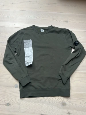 Olivgrön sweatshirt från CP company - Olivgrön sweatshirt från CP company med rund hals och ribbade muddar. Klassisk CP company-logga på vänster ärm. Tröjan är gjord i mjukt bomullsmaterial och har en avslappnad passform som är perfekt för en chill look. Kontakta för fler frågor eller bilder, kan gå ned i pris vid snabb affär 🤩