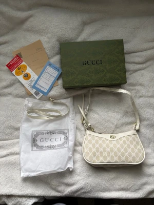 Gucci beige axelväska med GG-mönster - Säljer en snygg beige axelväska från Gucci med klassiskt GG-mönster i canvas och detaljer i vitt läder. Väskan har guldfärgade metalldetaljer och justerbar axelrem. Kommer med original dustbag och box. Perfekt för dig som vill ha en stilren och lyxig accessoar.