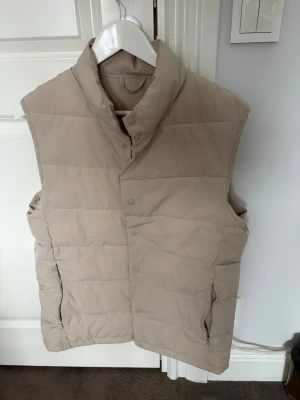 Beige dunväst med hög krage - Stilren beige dunväst med hög krage och tryckknappar framtill. Västen har två sidofickor med dragkedja och quiltad design för extra värme. Perfekt att slänga över en hoodie eller tröja för en chill look.