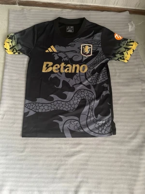 Svart fotbollströja med drakmotiv och Aston villa  - Cool svart fotbollströja från Adidas med stort grått drakmotiv på framsidan, gula detaljer och Betano-logga. Ärmarna har unikt mönster i gult och grönt samt tryckt klubbmärke på bröstet. Perfekt för dig som vill sticka ut på planen.
