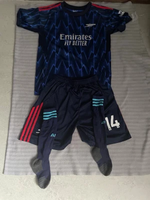 Arsenal mörkblå matchställ - Säljer ett mörkblått Arsenal matchställ med blixtmönster, bestående av tröja, shorts och strumpor. Tröjan har röda detaljer på axlarna och logga samt sponsortryck på bröstet. Shortsen har röda och turkosa ränder samt nummer 14. Perfekt för fotbollsträning!