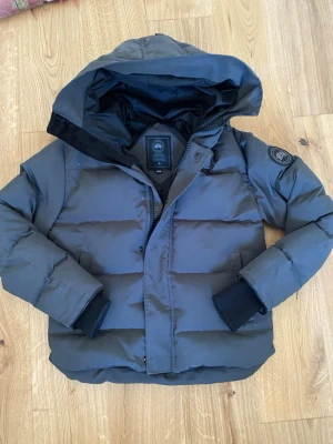 Mörkgrå pufferjacka från Canada Goose - Snygg mörkgrå pufferjacka från Canada Goose med stor huva och klassisk logotyp på ärmen. Jackan har breda kanaler, dolda muddar vid ärmsluten och dragkedja med knappslå. Perfekt för kalla vinterdagar och riktigt varm.