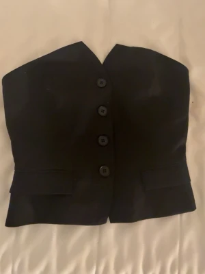 Svart bandeau väst från new yorker - Snygg svart bandeau väst från New Yorker med fyra knappar framtill och två dekorativa fickor. Västen är axelbandslös och har en stilren, croppad passform som passar perfekt till både jeans och kjol.