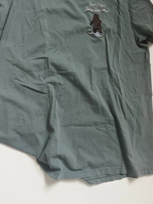 Grön t-shirt Snow Lodge brodyr - Säljer en grön t-shirt från Snow Lodge med broderad björn och texten 'Snow Lodge Greater Yellowstone' på bröstet. T-shirten har korta ärmar och är gjord i mjuk bomull. Perfekt för dig som gillar naturinspirerade plagg.