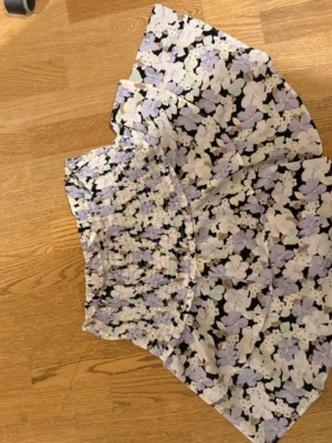 Blommig kortkjol med volang - Söt kortkjol med volang och blommigt mönster i lila, vit och svart. Kjolen har en smockad midja som ger en bekväm passform och ett flowy fall. Perfekt för dig som gillar en feminin och trendig stil.