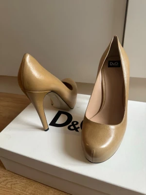 Beige pumps från D&G - Snygga beige pumps från D&G med rundad tå och hög smal klack. Skorna är tillverkade i glansigt skinn och har en klassisk, elegant design som passar perfekt till många olika outfits. Dold platå framtill för extra komfort. Klacken är 12 cm