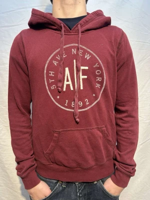 Vinröd hoodie från A&F med tryck - Snygg vinröd hoodie från Abercrombie & Fitch med stort rundat tryck på bröstet. Hoodien har huva med dragsko och en klassisk magficka. Perfekt för en avslappnad och trendig stil.