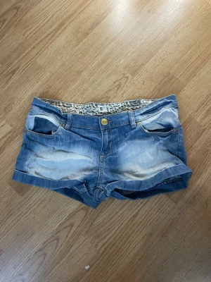 Blå jeansshorts med leoparddetalj - Snygga blå jeansshorts med slitningar och uppvikta ben. Insidan av linningen har ett leopardmönster som ger en cool detalj. Klassisk femficksmodell med knapp och dragkedja framtill. Perfekta för varma dagar! Lite högre pris då dom inte går att köpa längre men priset går att diskutera