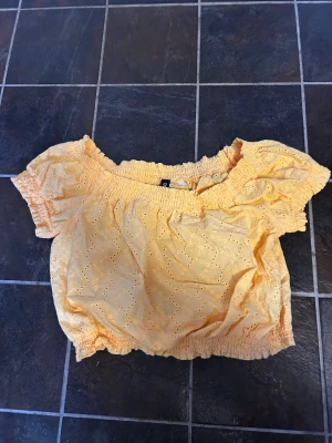 Gul broderad blus från H&M Divided - Söt gul blus från H&M Divided med broderade hål och puffiga korta ärmar. Modellen är croppad med resår vid halsringning och midja, vilket ger en avslappnad och trendig look. Perfekt för soliga dagar och sommarvibbar!