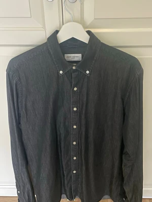 Svartgrå skjorta från John Henric - Stilren svart skjorta från John Henric med klassisk krage och vita knappar. Skjortan har lång ärm och är tillverkad i ett mjukt bomullstyg som passar perfekt till många olika looks. Varan är knappt använd. Storleken är M/L.