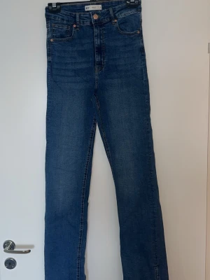 Blå raka jeans med slits från Gina Tricot - Snygga blå jeans från Gina Tricot med raka ben och hög midja. Jeansen har en cool slits längst ner på benen och klassiska fem fickor. Tillverkade i denim med en tidlös tvätt och kopparfärgade nitar. Perfekta för en trendig vardagslook.